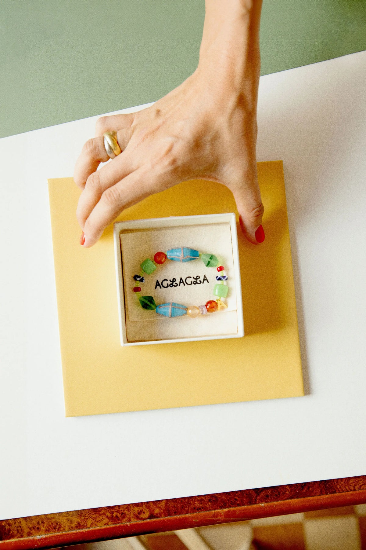 A hand with red nail polish and a gold ring holds a small white jewelry box containing a colorful beaded bracelet with the name ‘AGLAGLA’ written on it, set against a yellow card on a white table.
